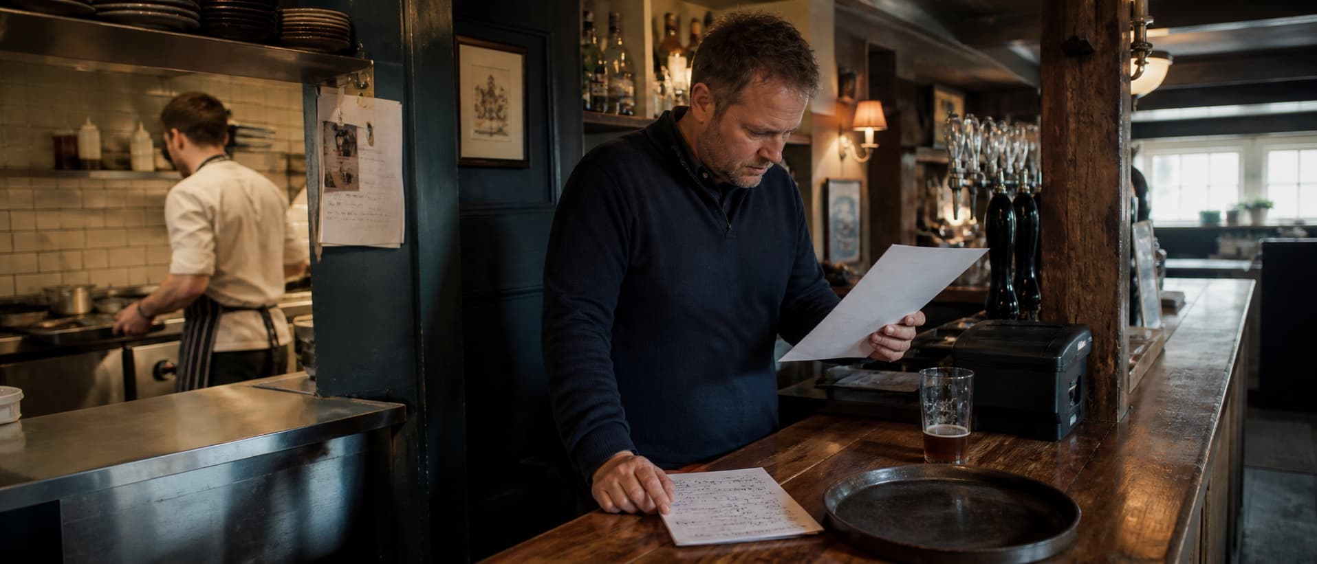 A pub manager checking supplier paperwork before service while a chef works in the kitchen